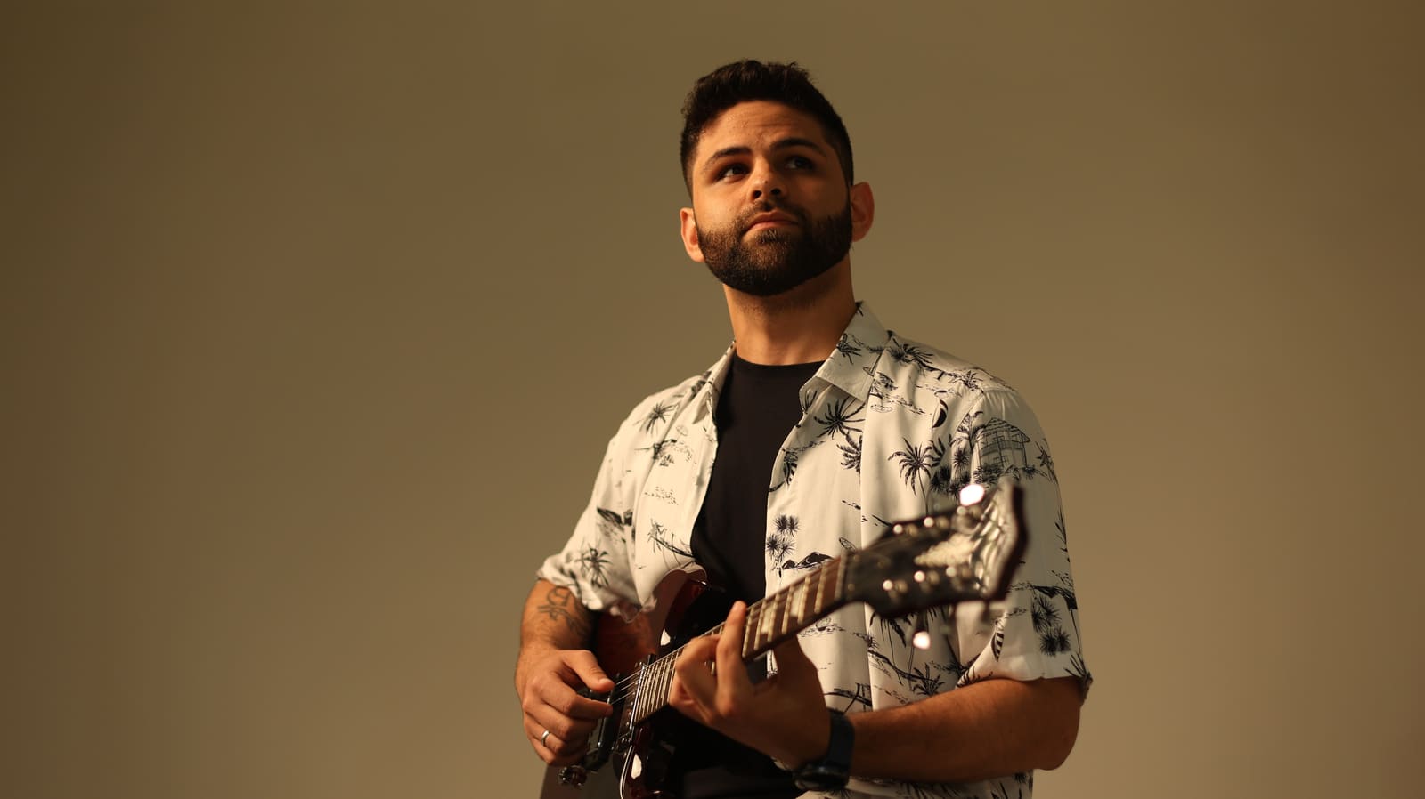 AQUINO com camisa estampada de palmeiras e guitarra em fundo bege
